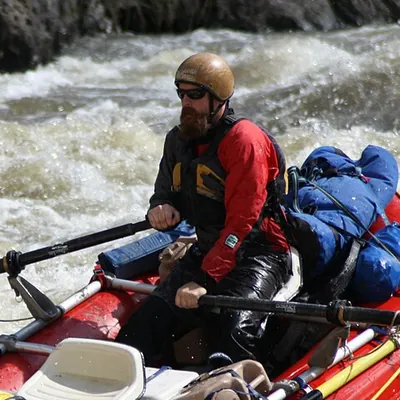 Marcus in a helmet and red jacket rowing a loaded raft through white water