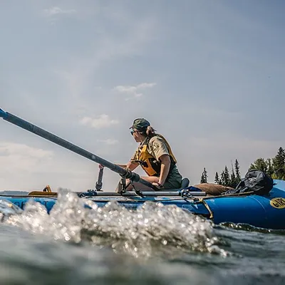 Jordan in a life jacket pulling the oars of a blue raft on a calm river